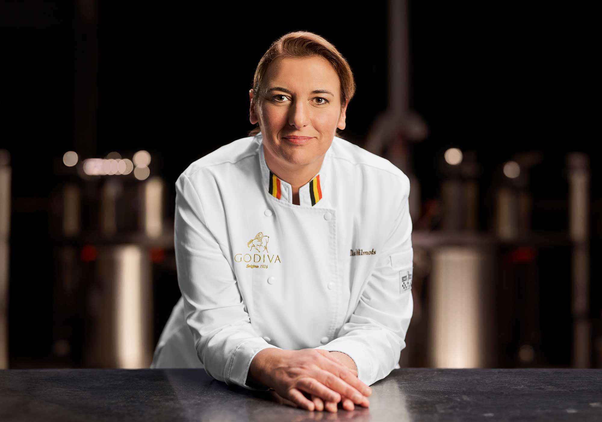 Chef Ilse Wilmots standing proudly in a kitchen with her GODIVA Chef's uniform on.
