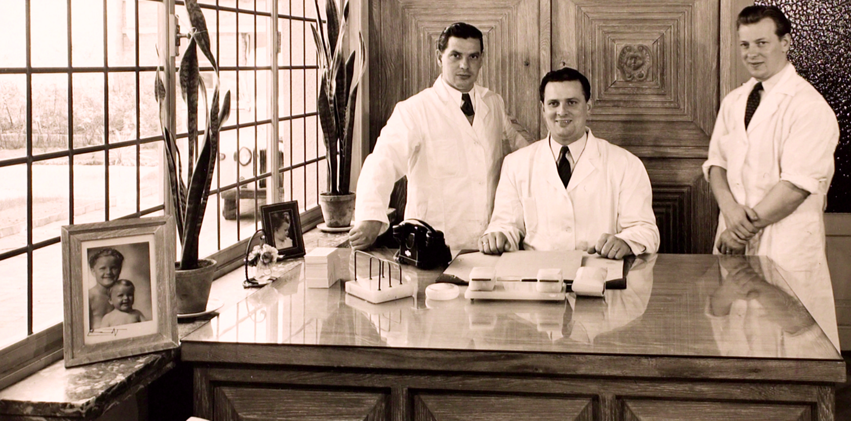 Three men in white lab coats sitting at a desk in a vintage office setting.