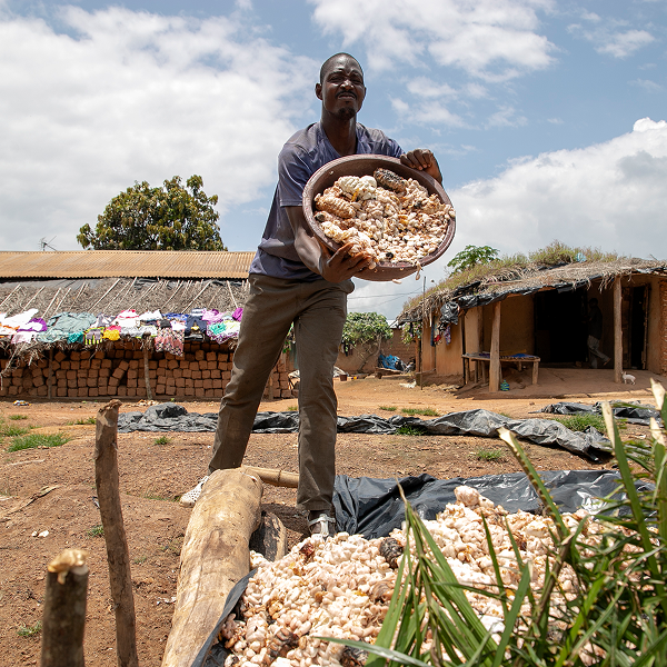 Man holding a bowl of food in an outdoor setting with huts and trees in the background